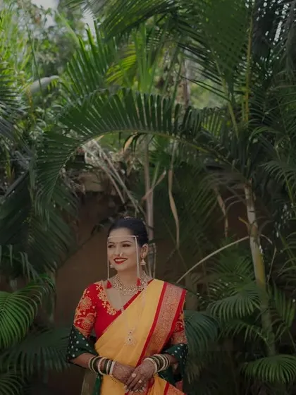 A radiant bride in her yellow and red saree, smiling against a backdrop of lush palm trees. A beautiful, natural bridal portrait.
