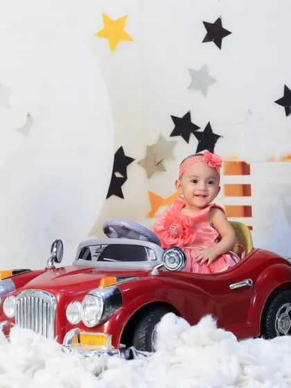 A baby girl in a pink dress enjoys a ride in a red toy car, set against our dreamy moon and stars backdrop.