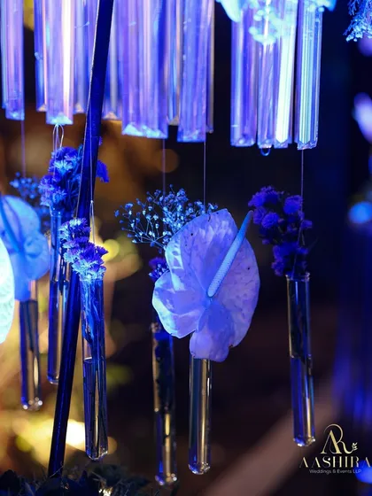 A close-up of the hanging test tube florals, showing a single anthurium bloom suspended in a glass vial, glowing under the blue light.