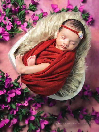 This classic newborn pose highlights the baby's delicate features. Swaddled in a rich red wrap and placed in a heart-shaped bowl surrounded by flowers, the composition creates a beautiful and artistic memory.