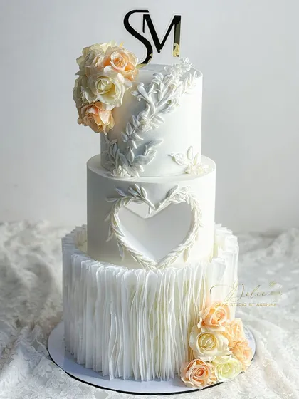 A wedding cake should be as beautiful as the love story it celebrates. This all-white, three-tiered fondant cake features delicate wafer paper ruffles, a heart-shaped cutout, and intricate leaf patterns. The soft peach and cream sugar flowers add a touch of timeless elegance.