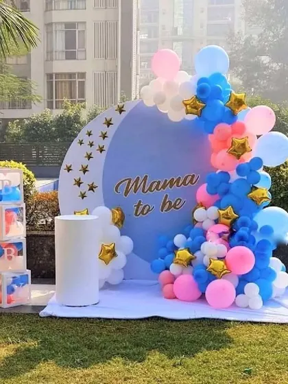 An outdoor 'Mama to be' baby shower setup, perfect for a garden or backyard party. The decoration includes a custom backdrop, a balloon garland in pink, blue, and white with star accents, and 'BABY' blocks.