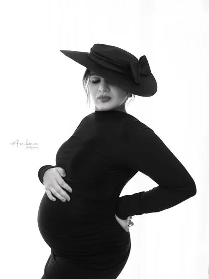 A black and white version of a fashion-forward portrait. The monochrome treatment adds a layer of drama and timelessness to the image, highlighting the textures of the dress and the bold shape of the hat.