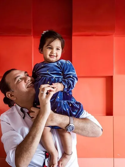 A father joyfully lifts his daughter onto his shoulders against a colorful, textured background. The image radiates paternal love and playful energy.