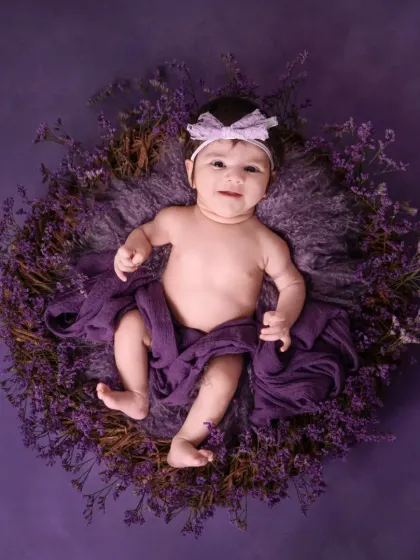 This adorable baby girl is nestled in a wreath of purple flowers against a solid purple backdrop. Her sweet smile and direct gaze make this a captivating infant portrait.