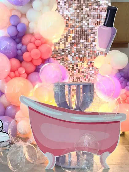 A focused shot of the spa party's central props, the pink bathtub and a nail polish cutout. The background features the shimmering sequin wall and a cascade of colorful balloons, emphasizing the fun, thematic elements.