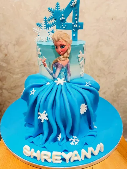 A magical two-tier Disney Frozen cake for a 4th birthday. This cake features an edible print of Elsa, whose dress is a beautifully draped fondant skirt, decorated with snowflakes.