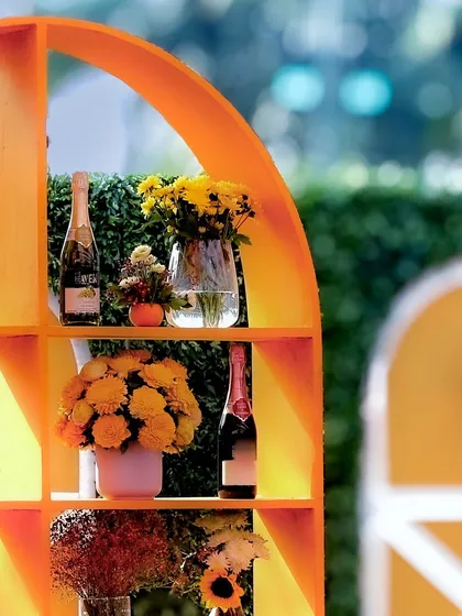 An arched shelving unit, painted bright orange, serves as a stylish bar back or display, decorated with champagne bottles and fresh flowers.