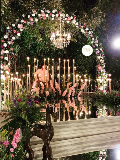 A stunning decor installation featuring a mirror reflecting a floral arch and a collection of tall, glowing candle stands. The wooden deer prop adds an enchanted element.