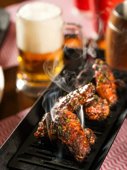 Smoky, grilled chicken wings on a black platter, with steam rising and a beer in the background. The action and moody lighting create a dynamic and appetizing shot for a pub menu.