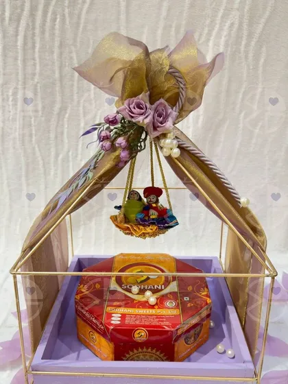 A beautiful Teej hamper featuring a Ghewar box placed inside a decorative metal cage adorned with a floral swing. The lavender color scheme adds a modern and elegant touch.