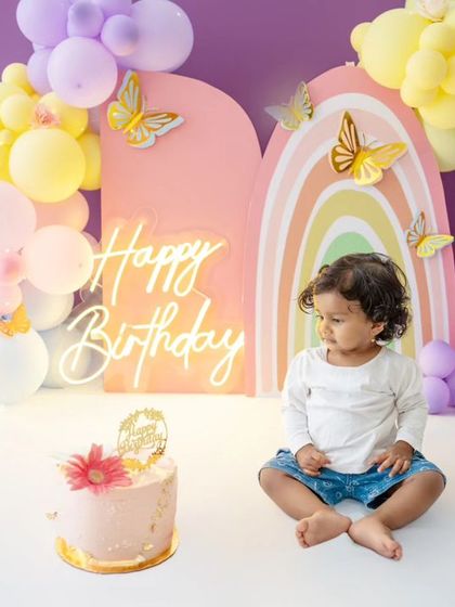The birthday girl sits patiently before her cake, with the beautiful, custom "Happy Birthday" neon sign and balloon arch in the background.