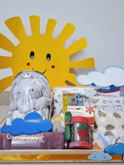 This 'Sunshine' themed hamper focuses on practical essentials. It includes diapers, wipes, a towel, a feeding container, and a soft elephant plush toy, all with a cheerful sun decoration.