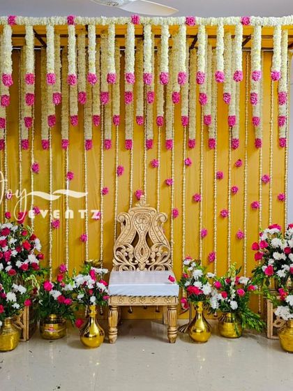 We designed this elegant stage with hanging garlands of tuberose and pink carnations against a bright yellow backdrop. The setup includes a golden chair and matching brass flower vases for a traditional ceremony.