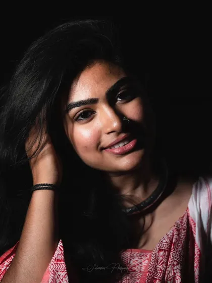 A high-contrast studio portrait. The makeup is designed to catch the light, with a focus on glowing skin and a bright smile that stands out against the dark backdrop.