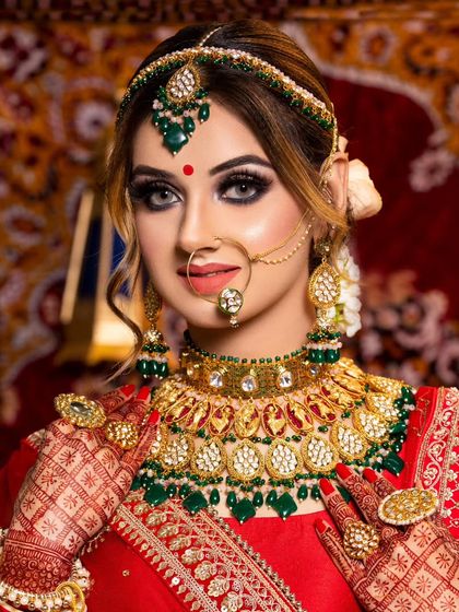 A smiling bride in a bright red lehenga with a heavy Polki and green bead jewellery set. The oversized rings and detailed matha patti add to the grandeur.