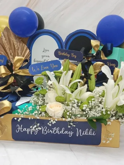 A sophisticated birthday hamper in blue, black, and gold. This crate features a fresh arrangement of white lilies and is filled with curated gifts for the birthday boy.