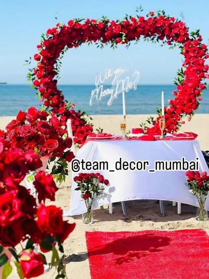 I design unforgettable marriage proposal setups, like this one on a Mumbai beach. It features a heart-shaped arch of red roses, a private dining table, and a 'Will you Marry Me?' sign, creating a picture perfect moment against the ocean backdrop.