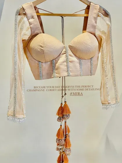 This custom champagne-colored corset blouse is for the modern bride. It features delicate silver detailing and sheer sleeves, perfect for pairing with a lehenga or saree for a reception or sangeet look.