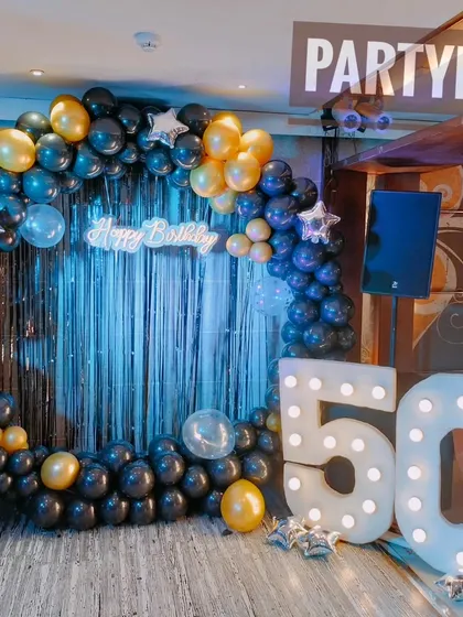 A 50th birthday party with a blue and gold balloon arch against a sequin backdrop, complete with large marquee numbers.