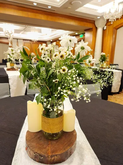 A rustic and charming centerpiece with white daisies and baby's breath in a burlap-wrapped mason jar. The arrangement is set on a wooden slice and surrounded by pillar candles.