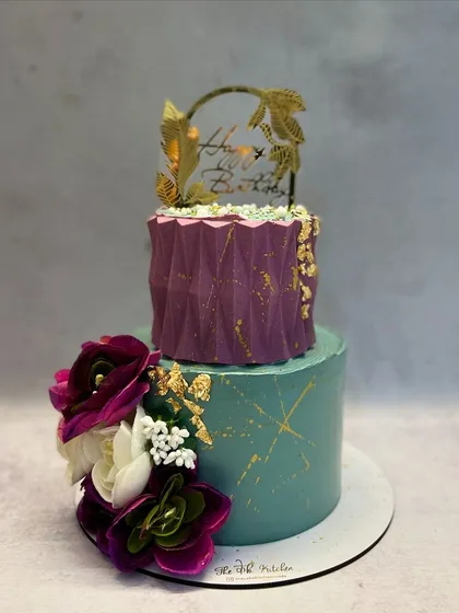 A modern two-tier cake combining a teal base with a textured purple top tier. The design is accented with gold leaf, abstract gold lines, and a beautiful arrangement of artificial purple and white flowers.