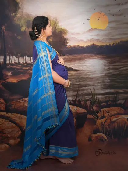 A beautiful studio portrait of a mother-to-be in a blue saree, set against a painted landscape backdrop with a full moon.