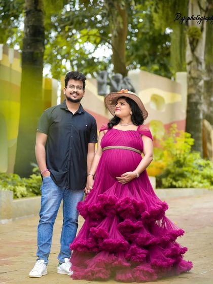 A sweet couple's portrait in a park-like setting. The mother-to-be is wearing a wine-colored ruffled gown and hat.
