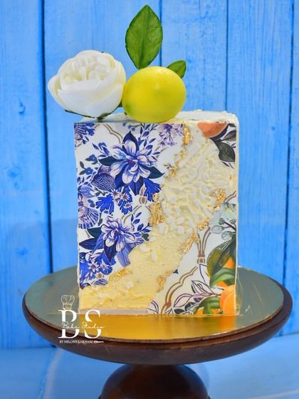 A premium square cake with an artistic, tile-like design. This elegant cake combines floral patterns with a textured white overlay and is topped with a fresh lemon and a white sugar flower.