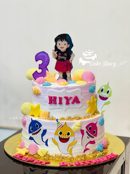 A fun-filled Baby Shark cake for Hiya's third birthday. This two-tier cake features the whole shark family swimming in a sea of pink and blue, surrounded by edible shells and sand.