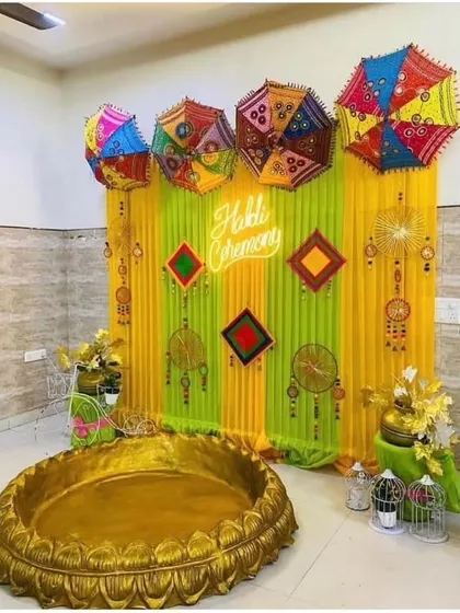 This Haldi ceremony setup features a neon sign, colorful umbrellas, and a large golden 'urli' for the main ritual. It's a mix of modern and traditional elements.