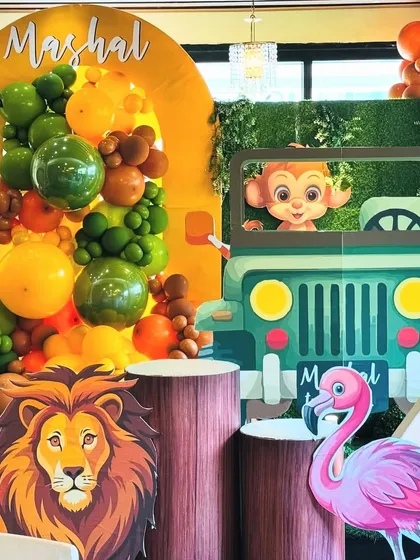 A detailed shot of the jungle party decor, focusing on the lion prop and the personalized jeep. The image highlights the rich textures, from the wood-patterned plinths to the green balloon garland, creating an authentic safari feel.