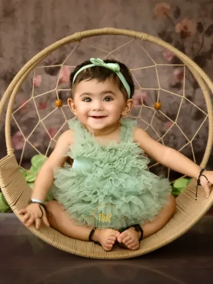 A happy baby girl in a green ruffled romper, sitting confidently in a round dreamcatcher swing.