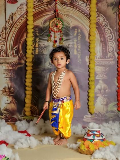 A handsome little Krishna. This full-length portrait shows off his adorable costume against a beautifully detailed temple-themed backdrop.
