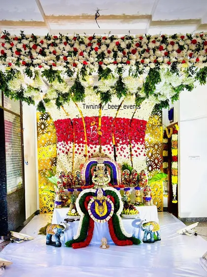 An elaborate floral mantap designed for a Varamahalakshmi puja at home. This decoration features a dense ceiling of white and red flowers flowing down into a multi-colored floral backdrop for the deity, creating a truly divine atmosphere.