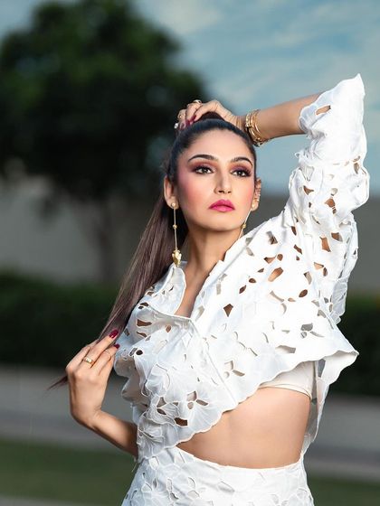 A high-fashion outdoor portrait for a magazine. The pose and high ponytail hairstyle complement the structured, modern design of the white laser-cut outfit.