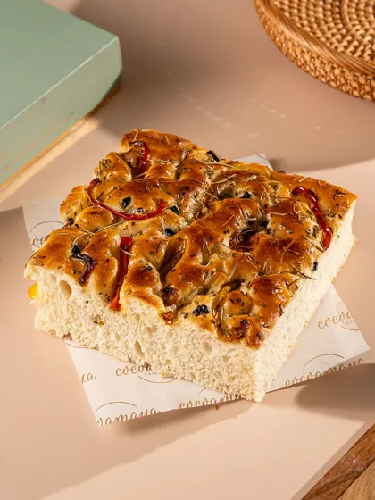 A thick, airy slice of focaccia bread with olives and herbs. The close-up shot makes it look soft and flavorful.