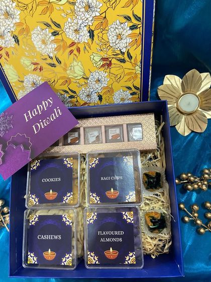 A beautiful floral-print box opens to reveal a neatly organized Diwali gift set. It includes four acrylic boxes with cashews, almonds, ragi chips, and cookies, alongside assorted chocolates.