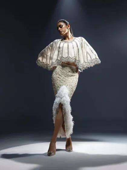 A full-length shot from the Kavita Arora Couture campaign, this image captures a dramatic pose under a spotlight. The styling highlights the garment's unique silhouette, with a heavily embellished cape top and a feathered, high-slit skirt.
