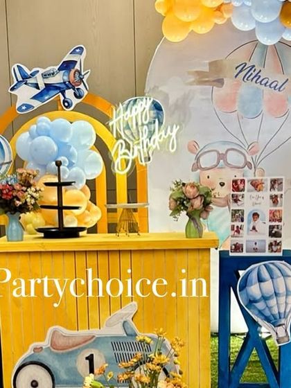 A hot air balloon and vintage vehicle theme for a first birthday. The setup features a bright yellow counter, a light-up number '1', and various props like airplanes and scooters.
