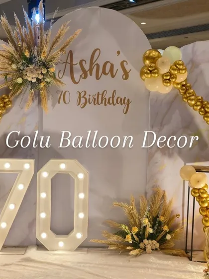 The main photo wall for a 70th birthday celebration. The setup includes the "Asha's 70 Birthday" custom text, marquee numbers, and floral accents with pampas grass.