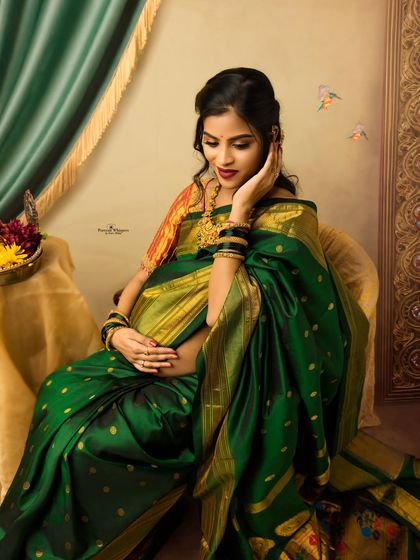 A delicate and thoughtful pose from a traditional maternity session. The mother-to-be, adorned in a green silk saree and gold jewelry, gently cradles her twin baby bump.