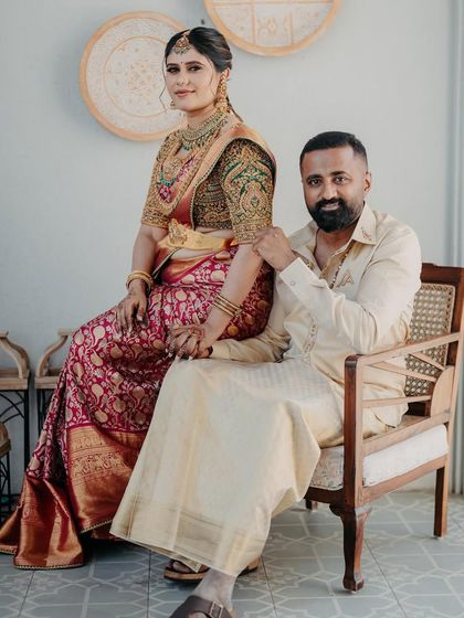A seated portrait of a couple. Her magnificent red and green bridal saree is the centerpiece, while his understated cream outfit provides the perfect complement.