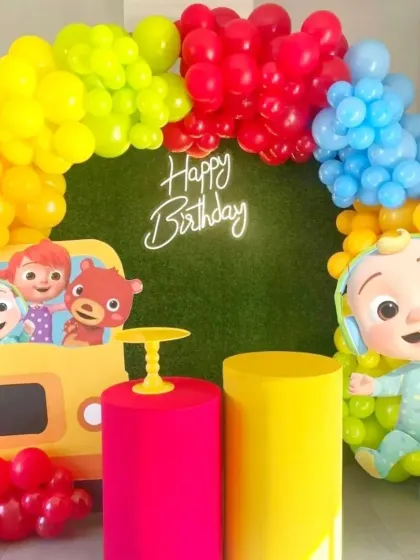 A vibrant and colorful Cocomelon-themed birthday party. The decor features a bright balloon arch over a faux grass wall, with large cutouts of JJ and other characters from the show.
