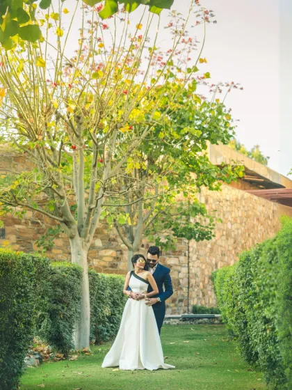 A romantic portrait of a couple in a beautiful garden setting. The groom embraces the bride from behind, framed by greenery, creating a classic and timeless pre-wedding photo.