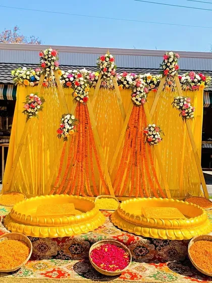 A classic outdoor Haldi setup with a bright yellow backdrop, marigold garlands, and traditional decorative umbrellas. The arrangement includes low seating and bowls of flower petals for the ceremony.