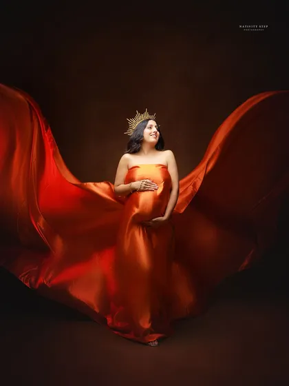 A regal portrait of the mother-to-be in a flowing red-orange gown, complete with a crown. The fabric toss creates a sense of power and majesty.