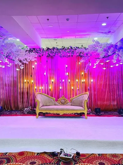 An elegant reception stage with a pink draped backdrop, illuminated by warm bulb lights and flanked by two white cherry blossom trees.