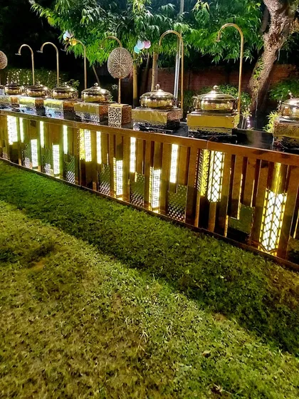 A beautifully lit rustic wooden buffet counter set up outdoors. The warm internal lighting and traditional brass serving dishes create a cozy and inviting atmosphere for an evening gathering.