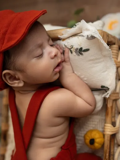 A newborn in a red cap and overalls, sleeping peacefully in a wicker bed. The composition, with white flowers hanging above, creates a beautiful, story-like image.